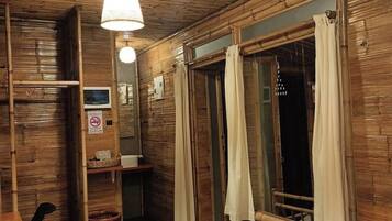A room with wooden walls and ceiling, a bed with a white duvet, a small desk with a chair, a mirror, and a window with curtains.
