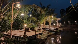 Una terrassa de fusta amb barana, envoltada de palmeres i una petita zona d'aigua, amb il·luminació ambiental.
