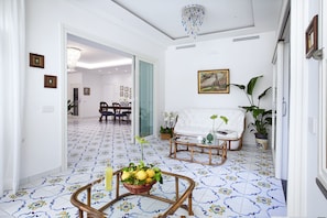 A spacious living room with a patterned tile floor, a white sofa, a coffee table with a basket of fruit, and a dining area in the background. - AMORE RENTALS - Villa Flavia with Private SPA, Swimming Pool, Terraces, Sea View (Sorrento)