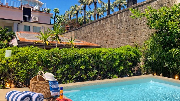 A pool area with a clear view of the pool, a towel, a hat, and a bottle, surrounded by greenery and a stone wall.
