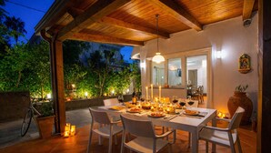 An outdoor dining area with a wooden pergola, white chairs, a table set for a meal, candles, and a bicycle parked nearby.