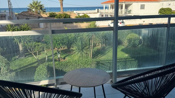 Un balcón con dos sillas negras y una mesita pequeña, con vistas a una zona costera con casas y palmeras.