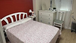 Un dormitorio con cama, mesita de noche, cómoda, silla, ventana con persianas y radiador.