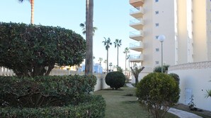 Un jardín cuidado con setos ordenados y un pequeño árbol, un edificio blanco con balcones y un cielo despejado.