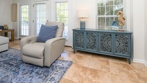 Un salon avec un canapé beige, un tapis à motifs, un meuble turquoise orné de sculptures décoratives et une fenêtre avec des stores.
