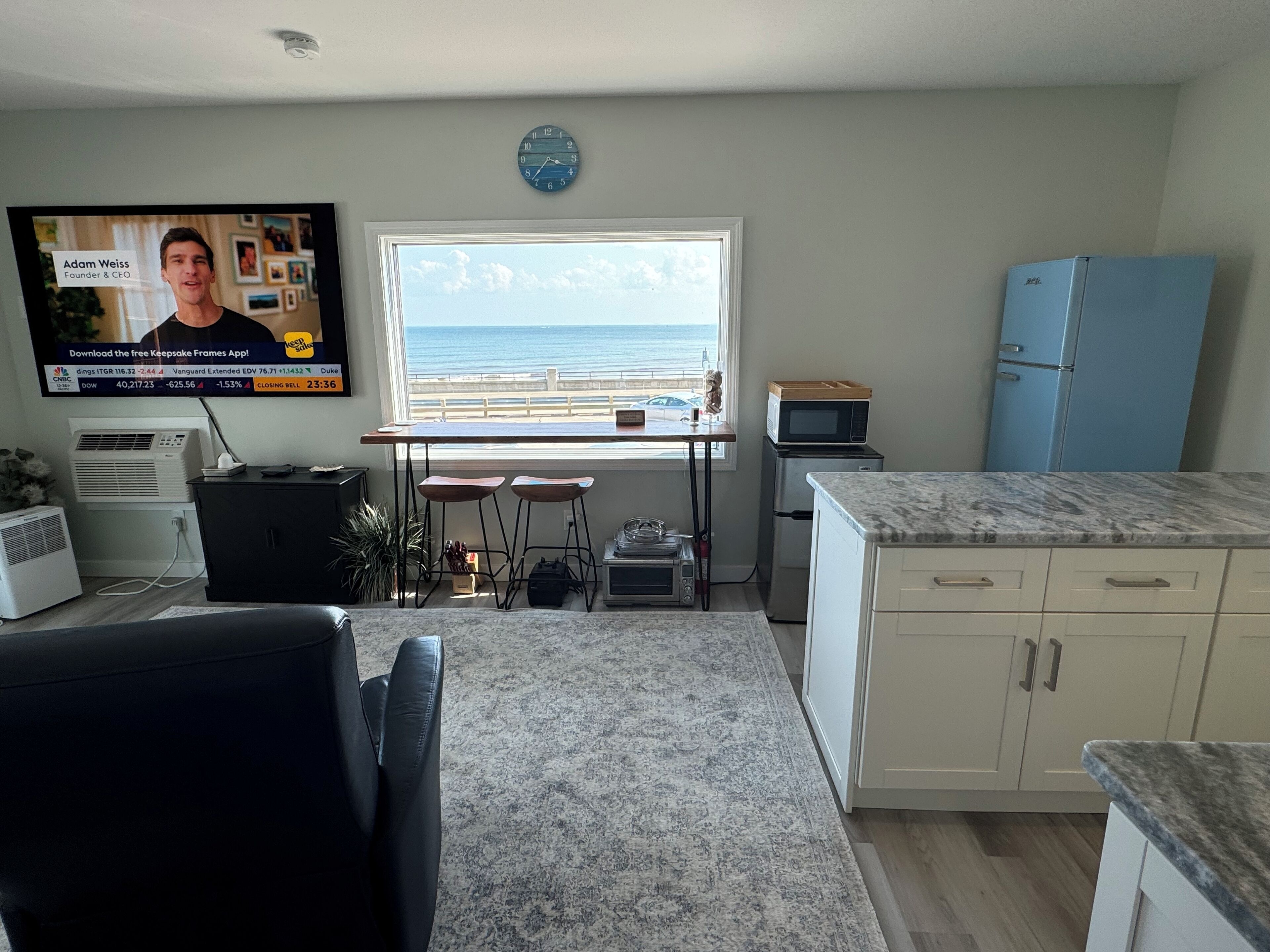 A modern kitchen with a view of the ocean, a dining area with a TV displaying a news program, and a small kitchenette.