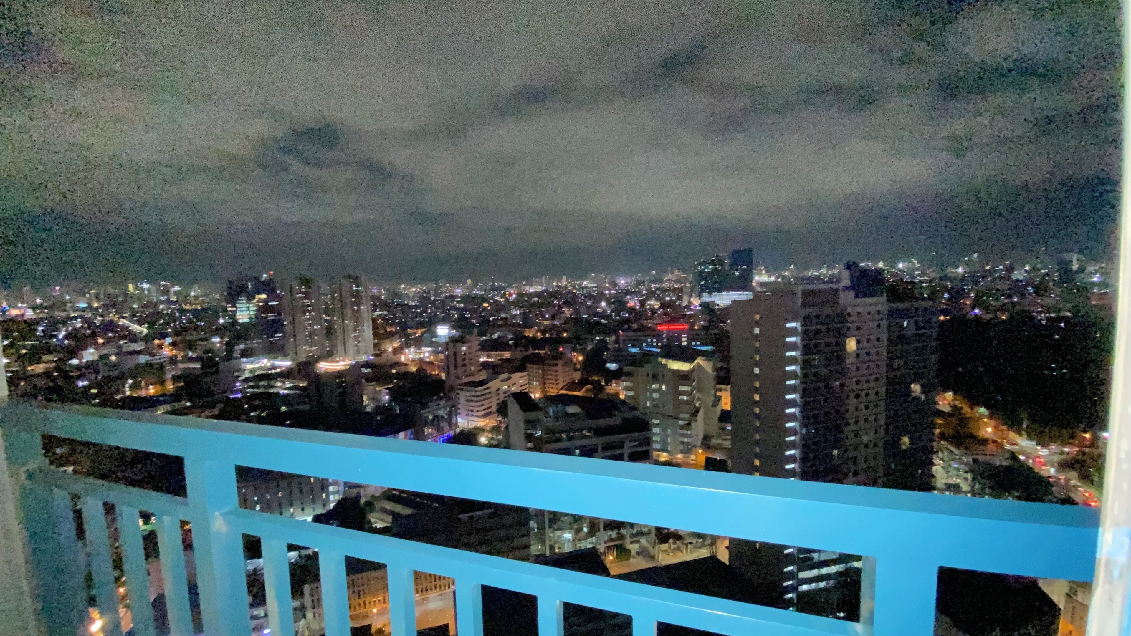 A nighttime cityscape viewed from a balcony with a railing.