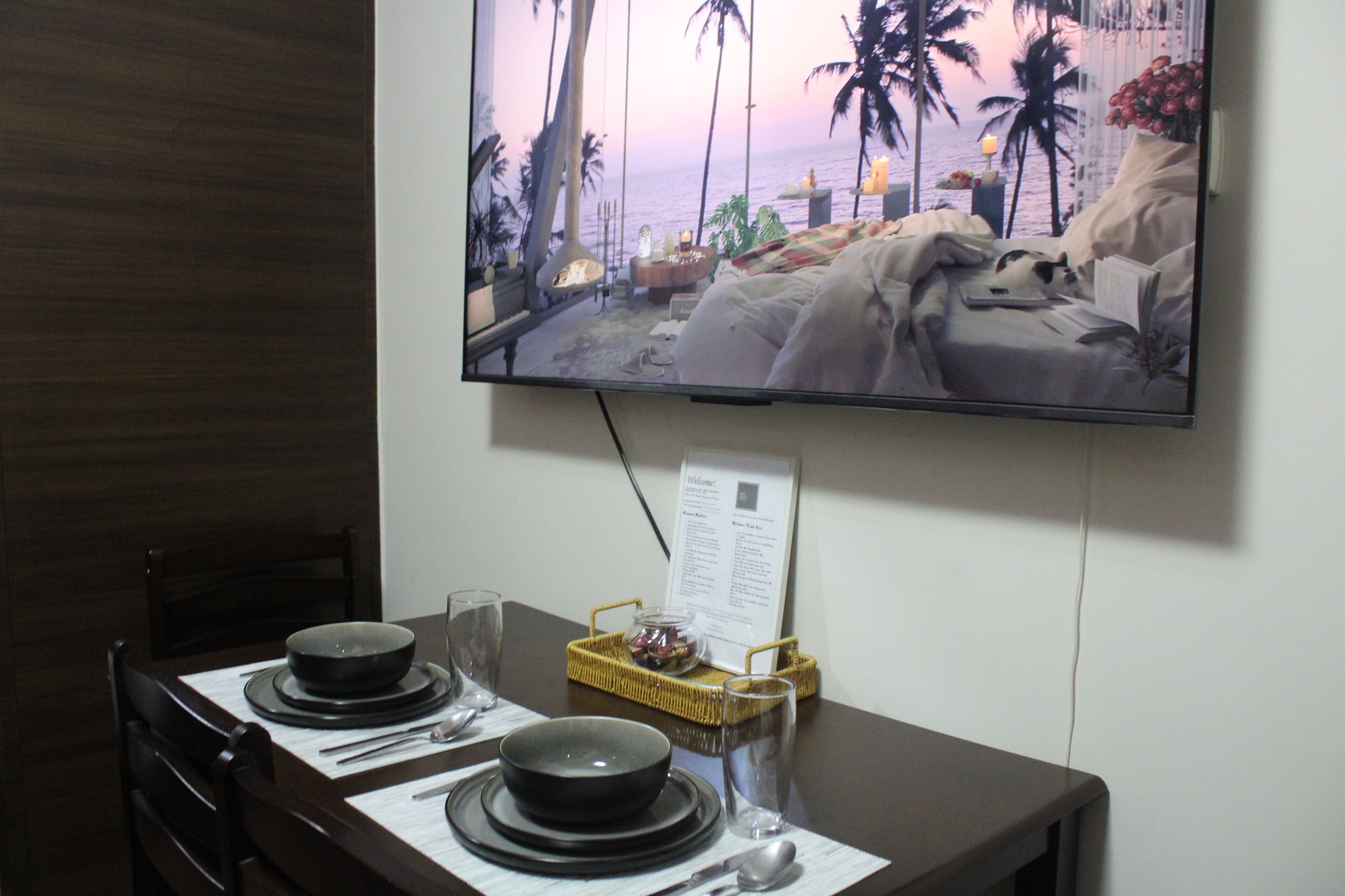 Un comedor con una mesa dispuesta para dos, un televisor en la pared mostrando una escena de playa y un atril para el menú.