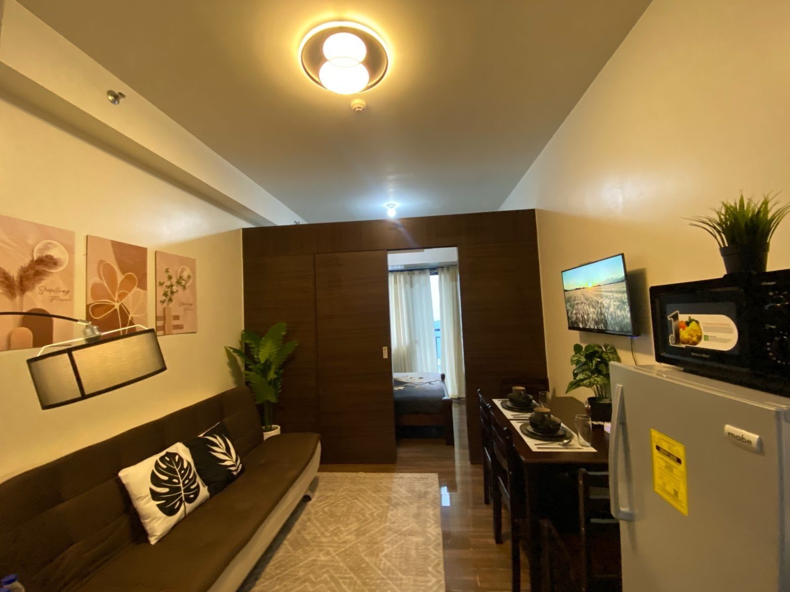 Ein modernes Wohnzimmer mit Sofa, Essbereich mit Tisch und Stühlen, einer Küche mit Kühlschrank und einem an der Wand montierten Fernseher.
