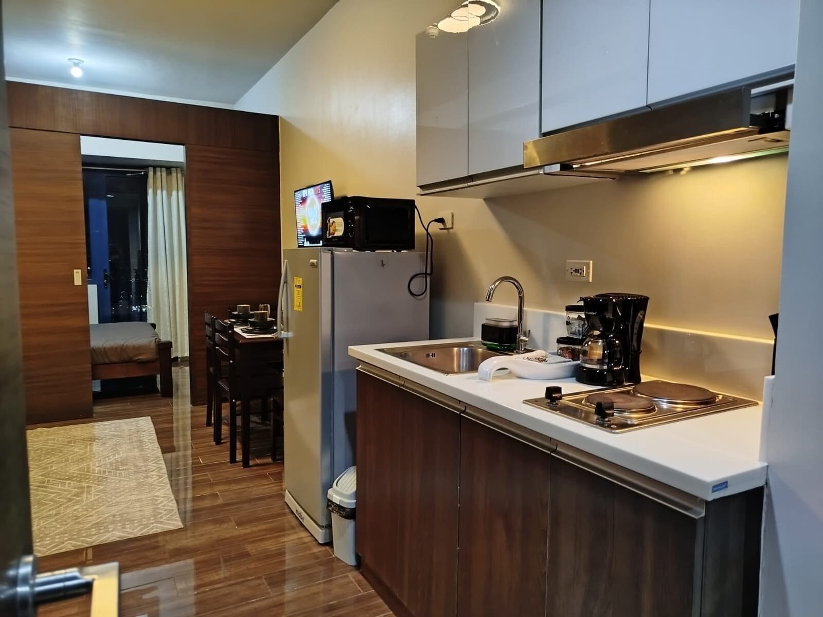 A compact kitchen with a refrigerator, microwave, sink, and coffee maker. There is a dining area with a table and chairs visible through an open doorway.