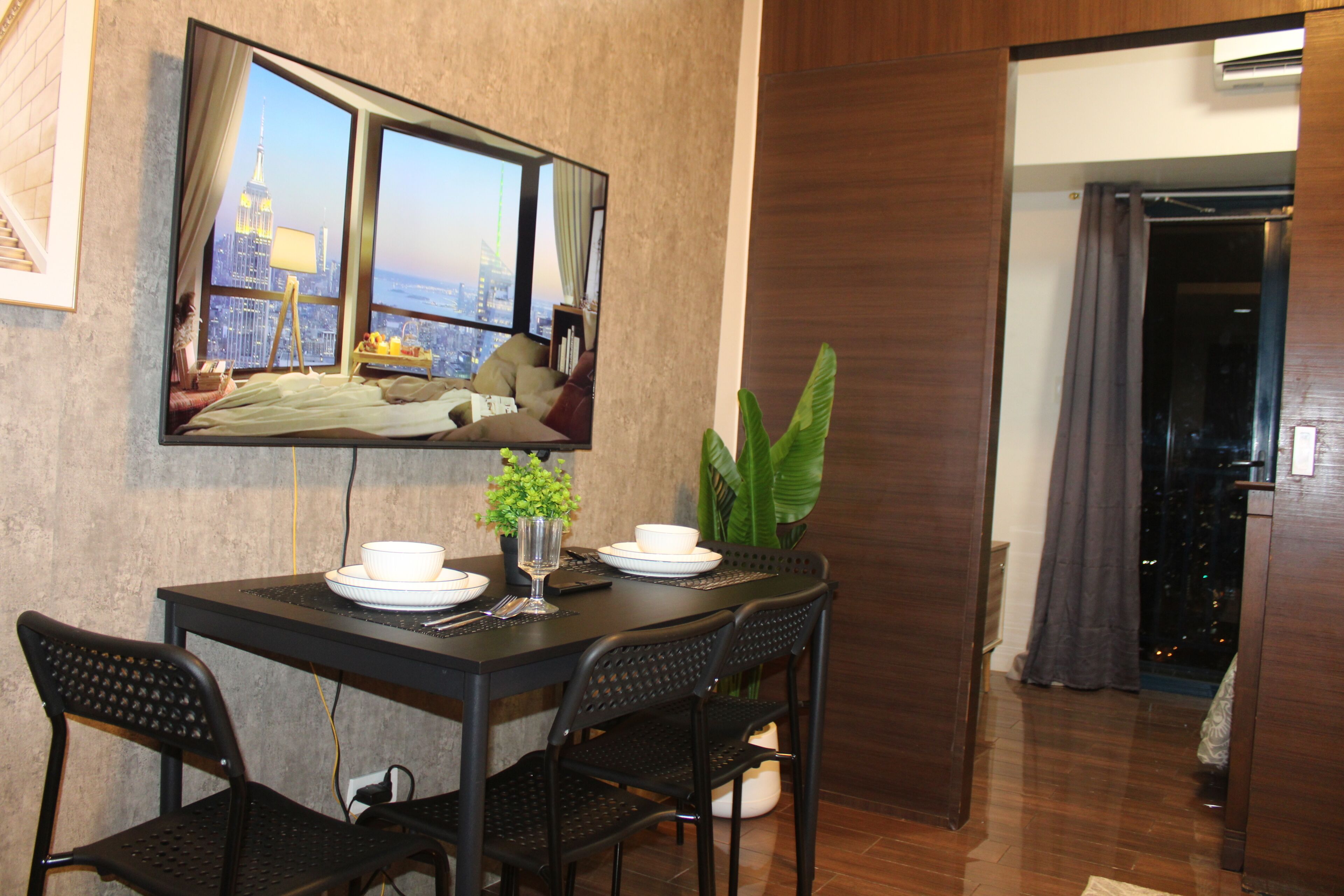 A dining area with a table set for two, a mirror reflecting a cityscape, and a potted plant.