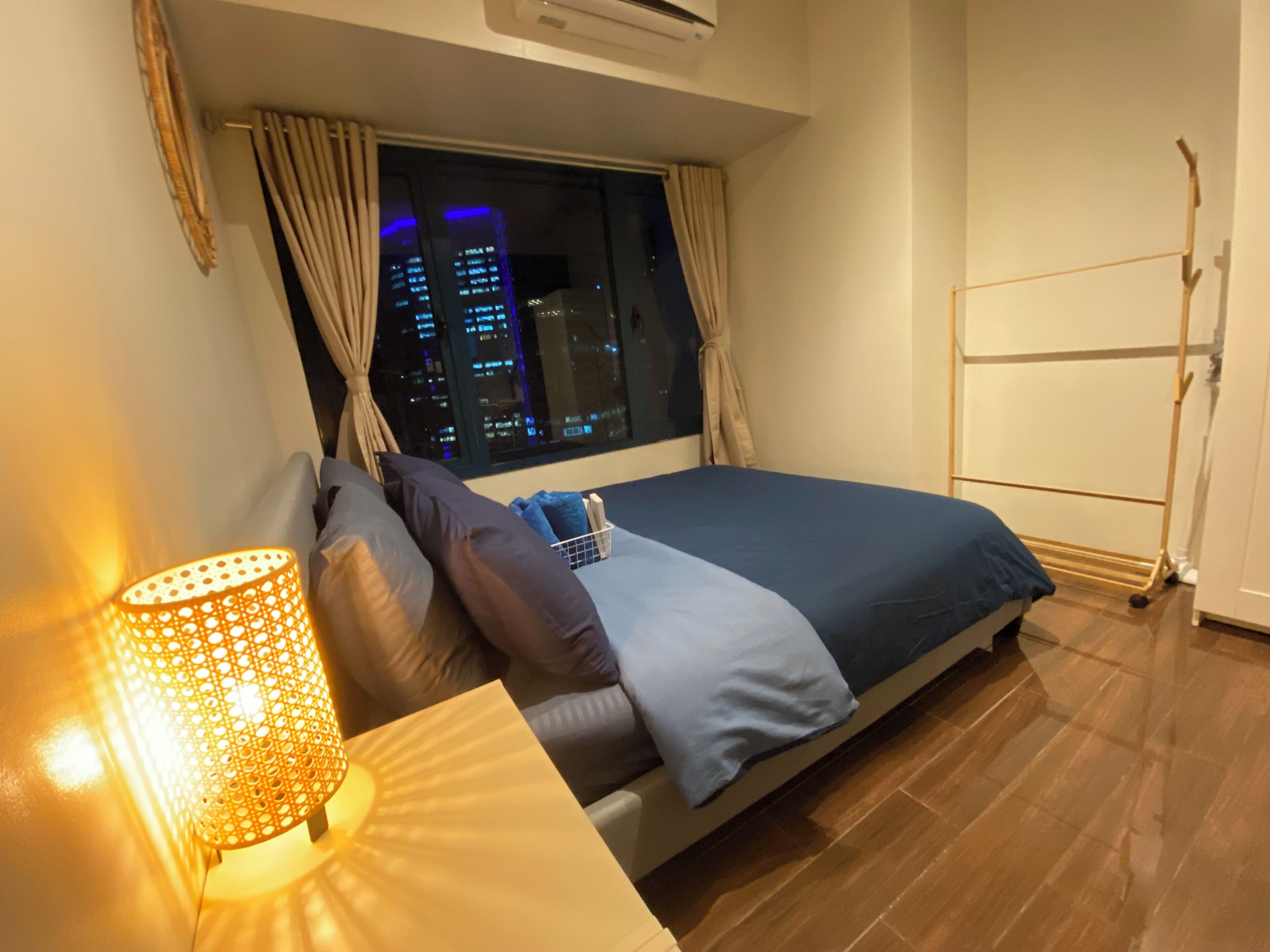 A bedroom with a bed, a nightstand, a lamp, a window with a city view, and a wardrobe.
