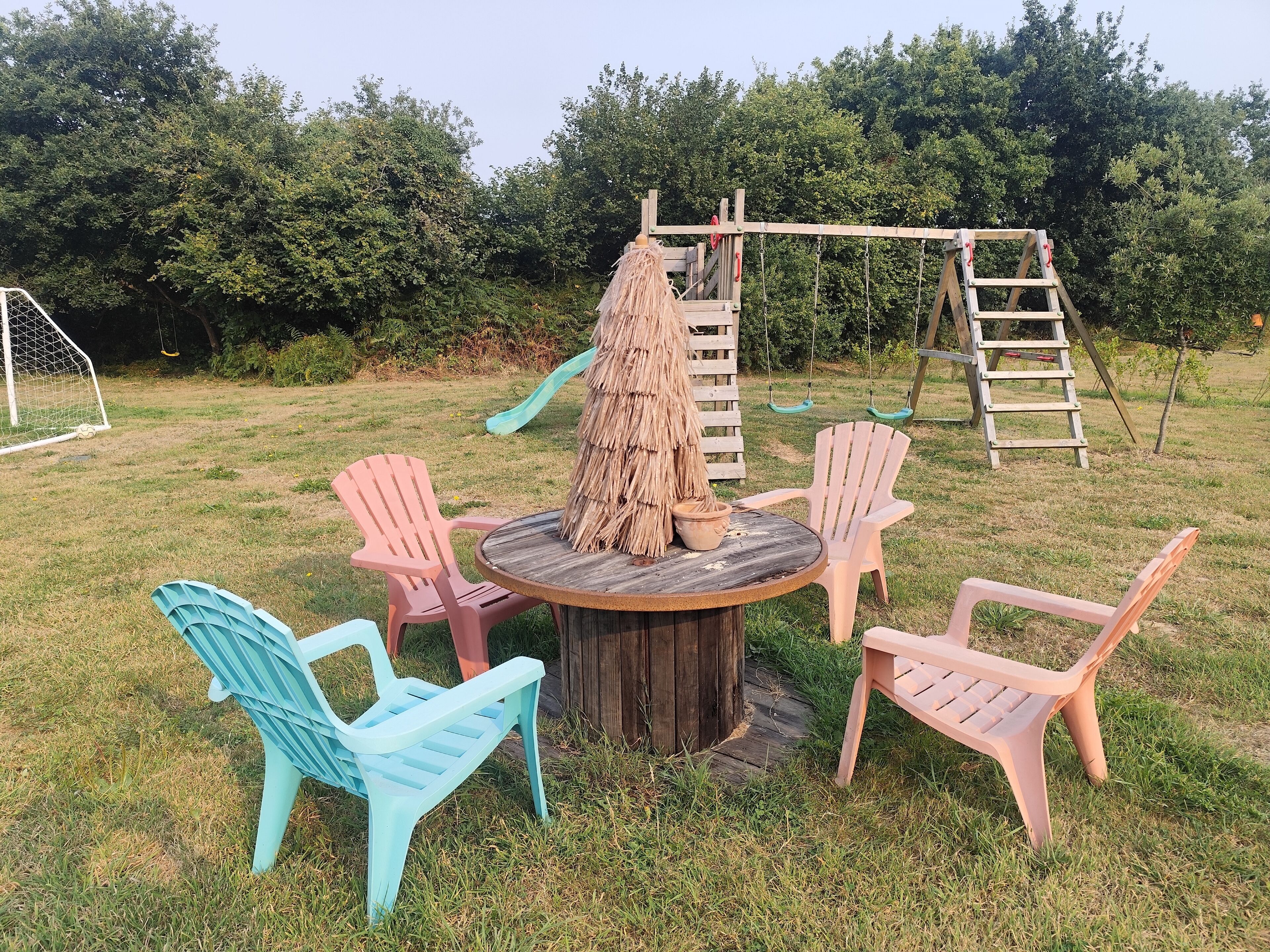 Una mesa redonda de madera rodeada por cuatro sillas, un toldo de paja y un columpio de madera en un espacio al aire libre cubierto de césped.