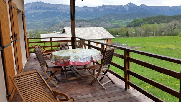 Una terraza de madera con una mesa y sillas, con vista a un paisaje verde y montañas a lo lejos.