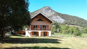 Un chalet de dos plantas con balcón, rodeado de un paisaje cubierto de hierba y una montaña al fondo.