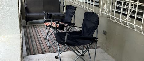 Ett takförsett utomhusområde med sittplatser, ett bord och stolar, en hopfällbar stol i svart och en grill av metall som hänger på väggen.