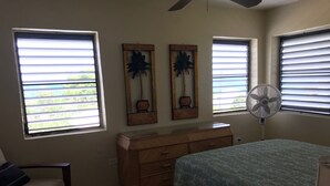 A bedroom with a bed, a dresser, two wall-mounted fans, and two windows with blinds. - Bananaquit House (Frederiksted)
