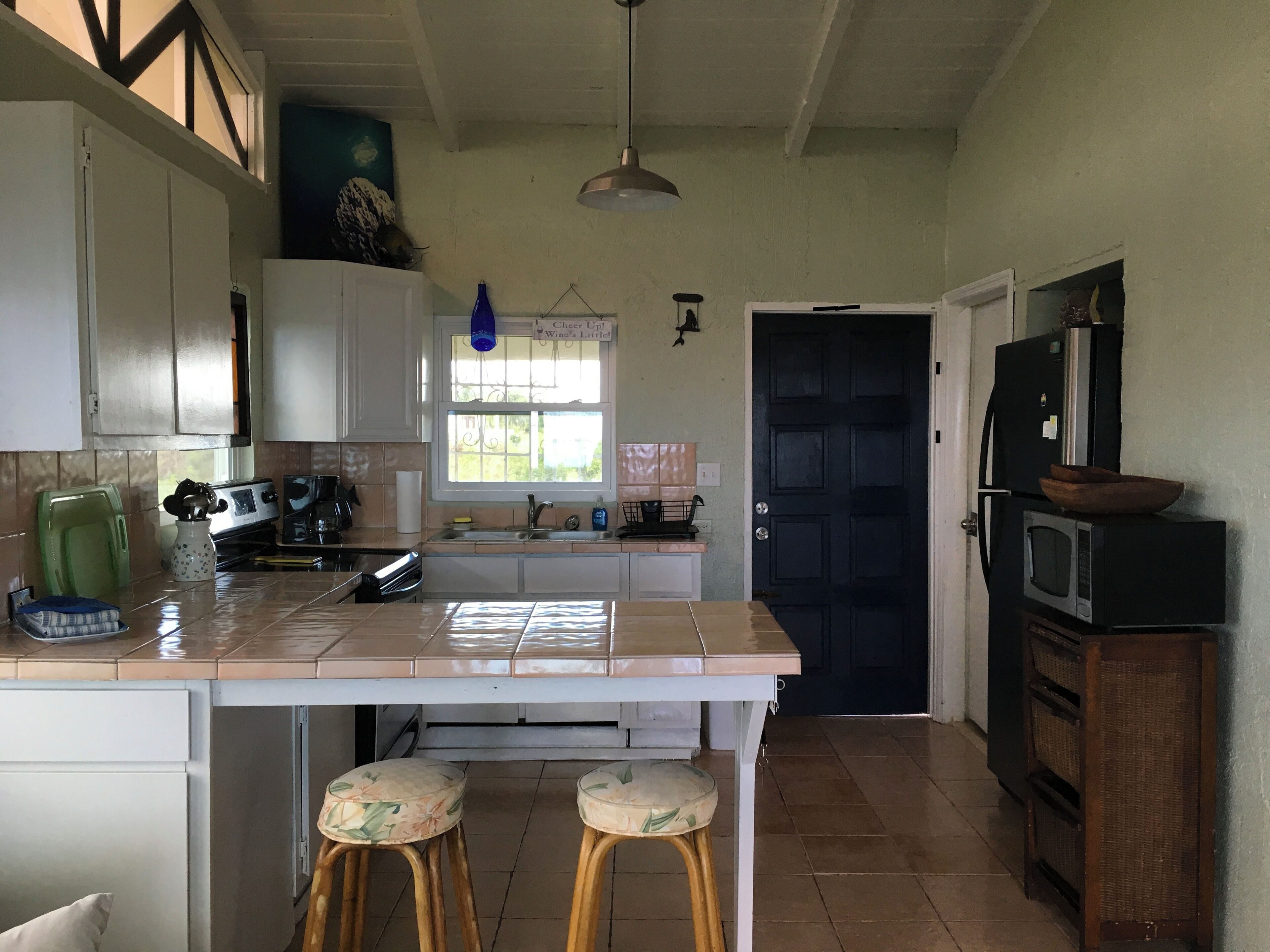 Une cuisine avec des meubles blancs, un pare-feu en carrelage, une fenêtre donnant sur un beau paysage, un évier, une cuisinière et deux tabourets.