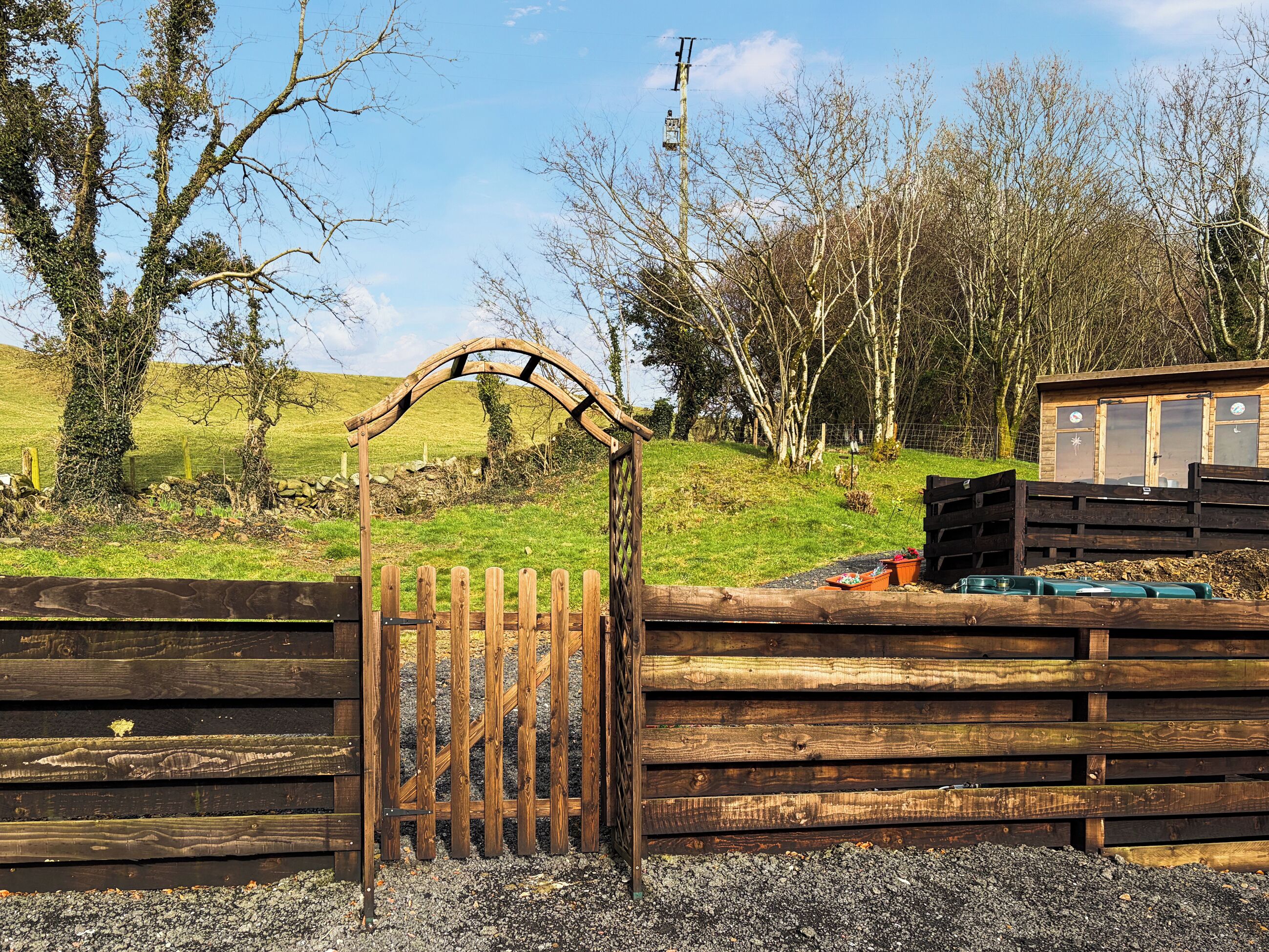 A wooden gate with a curved top, a fenced area with a wooden structure, and a grassy landscape with trees.