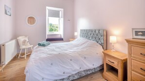 Un dormitorio con cama, mesita de noche con lámpara, silla, radiador y ventana con vistas.