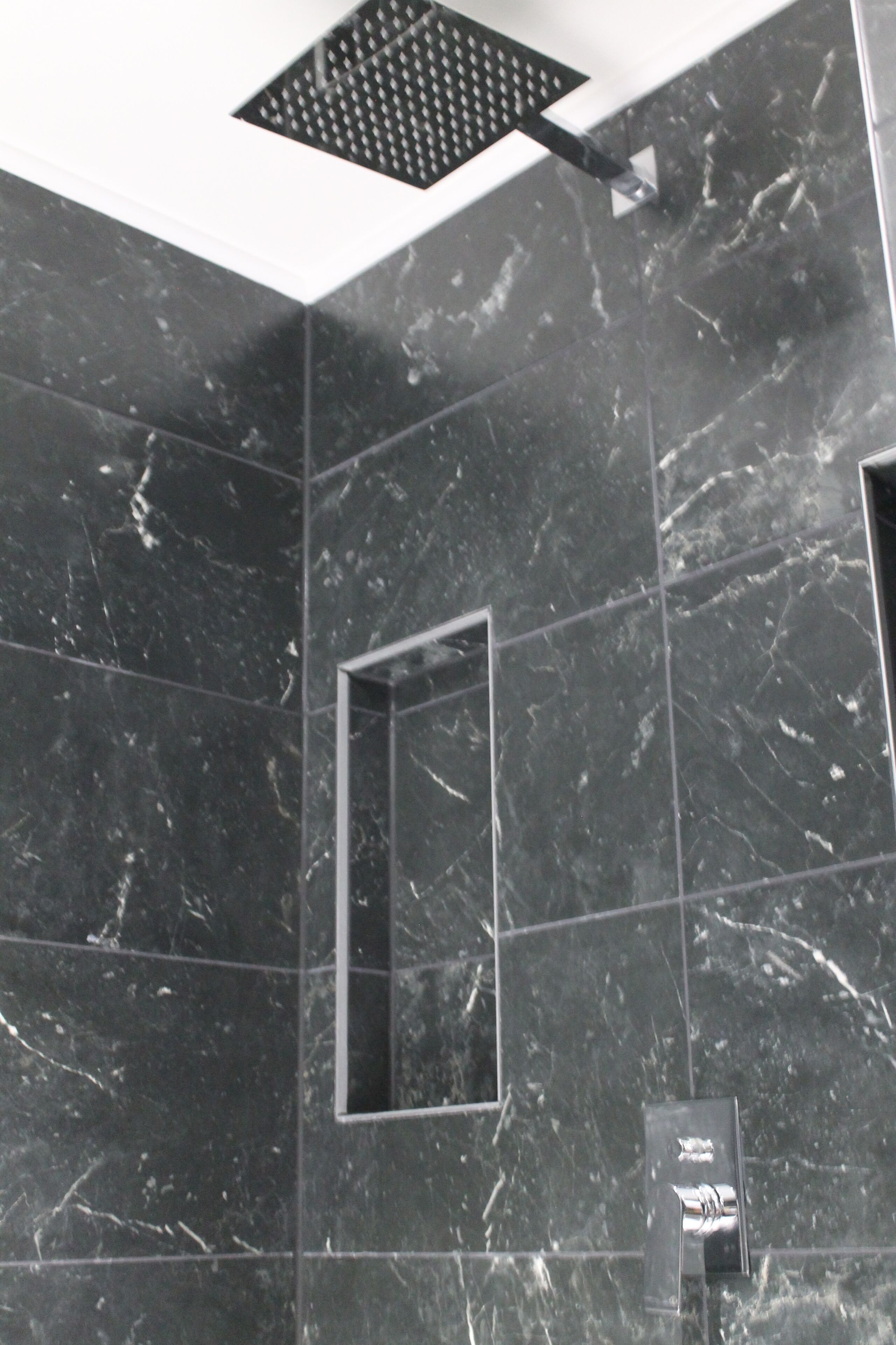 Une salle de bain moderne avec des carreaux de marbre foncé, une pomme de douche carrée et un miroir rectangulaire.