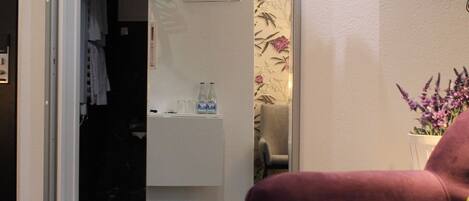 Une chambre d’hôtel moderne avec un fauteuil violet, un téléviseur à écran plat fixé au mur, une porte donnant sur une salle de bain et une petite table sur laquelle est posé un vase de fleurs.