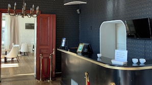 Zona de recepción de un hotel con mostrador, una araña de cristal y vista a un comedor con sillas y una mesa.