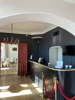 A hotel reception area with a reception desk, a chandelier, and a view of a dining area with chairs and a table.