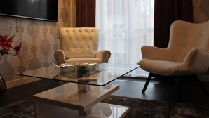 Una sala de estar moderna con una mesa de centro de vidrio, dos sillones blancos, un sofá, un televisor de pantalla plana y vistas a la ciudad a través de ventanas amplias.