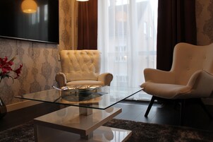 A modern living room with a glass coffee table, two white armchairs, a sofa, a flat-screen TV, and a view of the city through large windows.