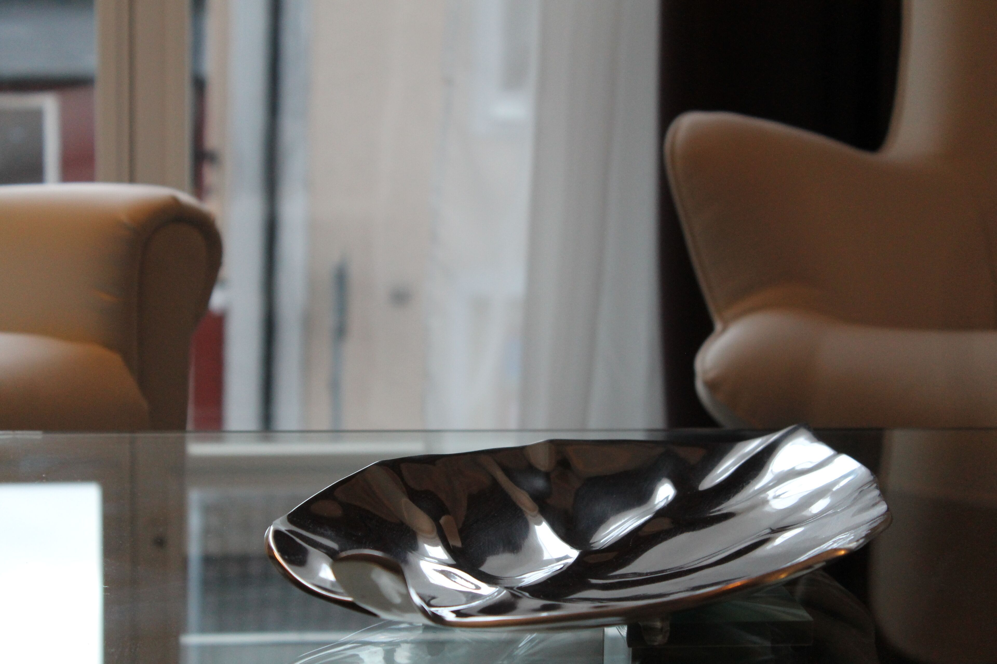 A modern interior with a reflective metallic bowl on a glass table, leather armchairs, and a window with blinds.