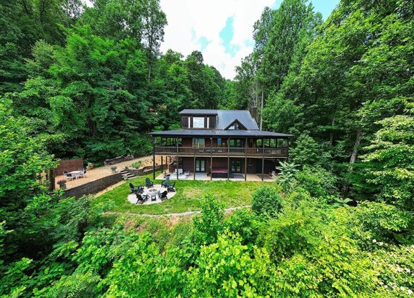 A two-story house with a deck and a fire pit in the backyard, surrounded by dense forest.