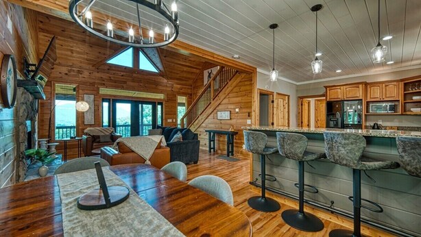 A spacious living area with a wooden ceiling, a dining table with chairs, a bar area with stools, and a kitchen with wooden cabinets. - Meribel-Hot tub-Fire pit-Amazing Views (Young Harris)