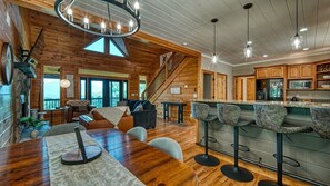 A spacious living area with a wooden ceiling, a dining table with chairs, a bar area with stools, and a kitchen with wooden cabinets.