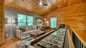 A cozy room with a wooden ceiling, a sofa, a coffee table, a lamp, and a large window with a view of trees.