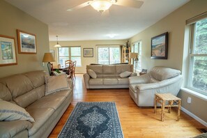 A spacious living room with a ceiling fan, large windows, and a wooden floor.