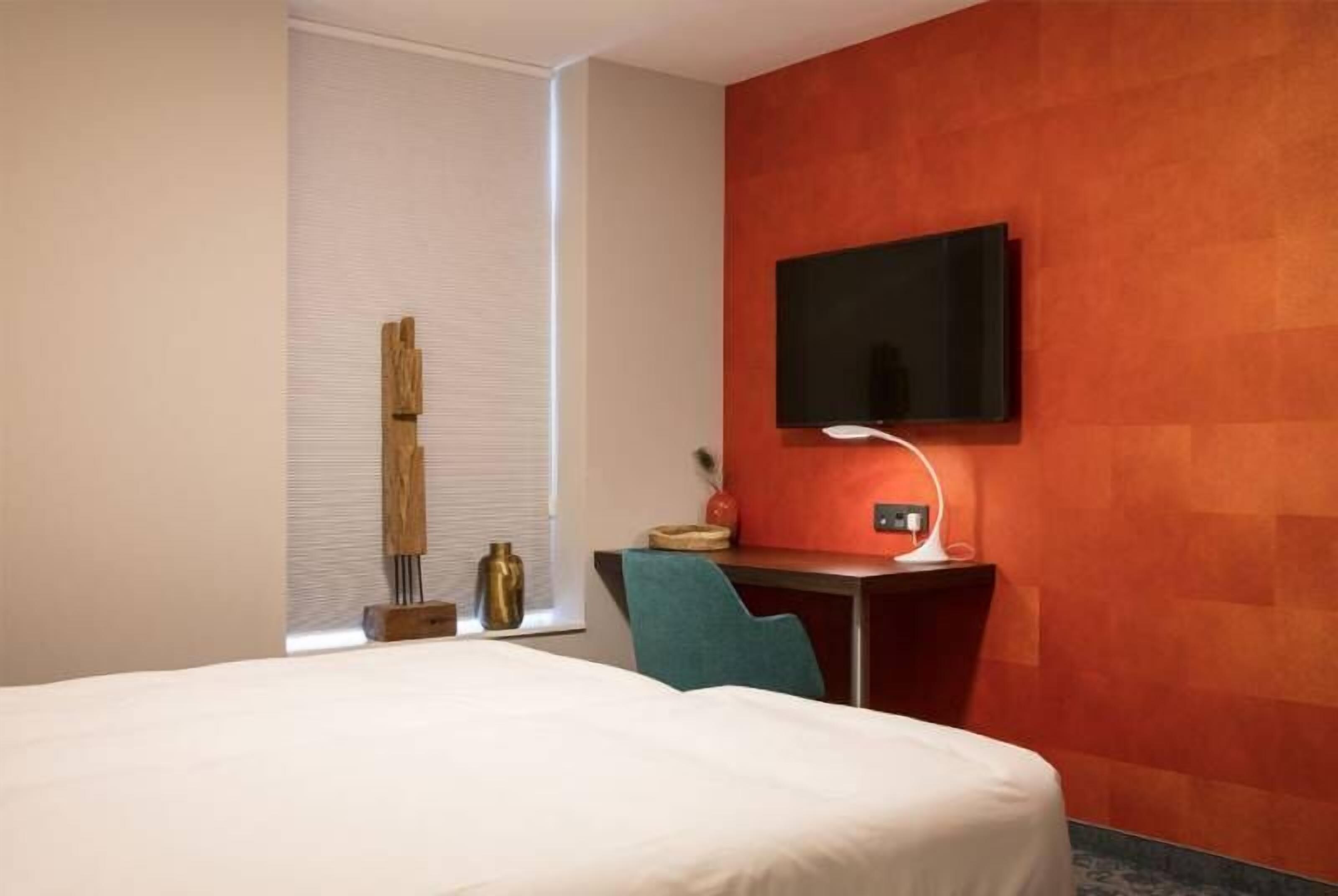 A hotel room with a bed, a desk with a TV, a lamp, and a chair.