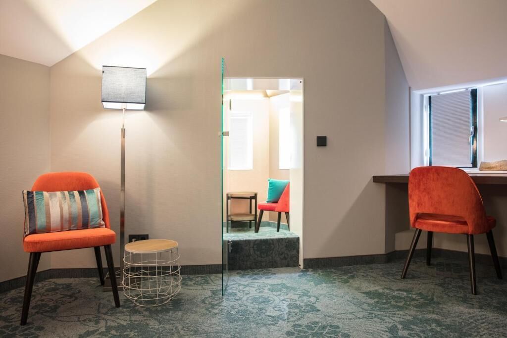 A room with a sloped ceiling, a floor lamp, two orange chairs, a small round table, and a view of a hallway.