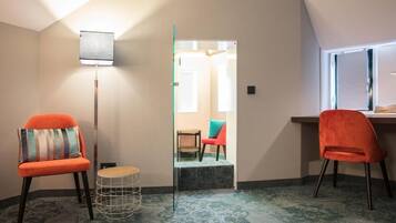 A room with a sloped ceiling, a floor lamp, two orange chairs, a small round table, and a view of a hallway.