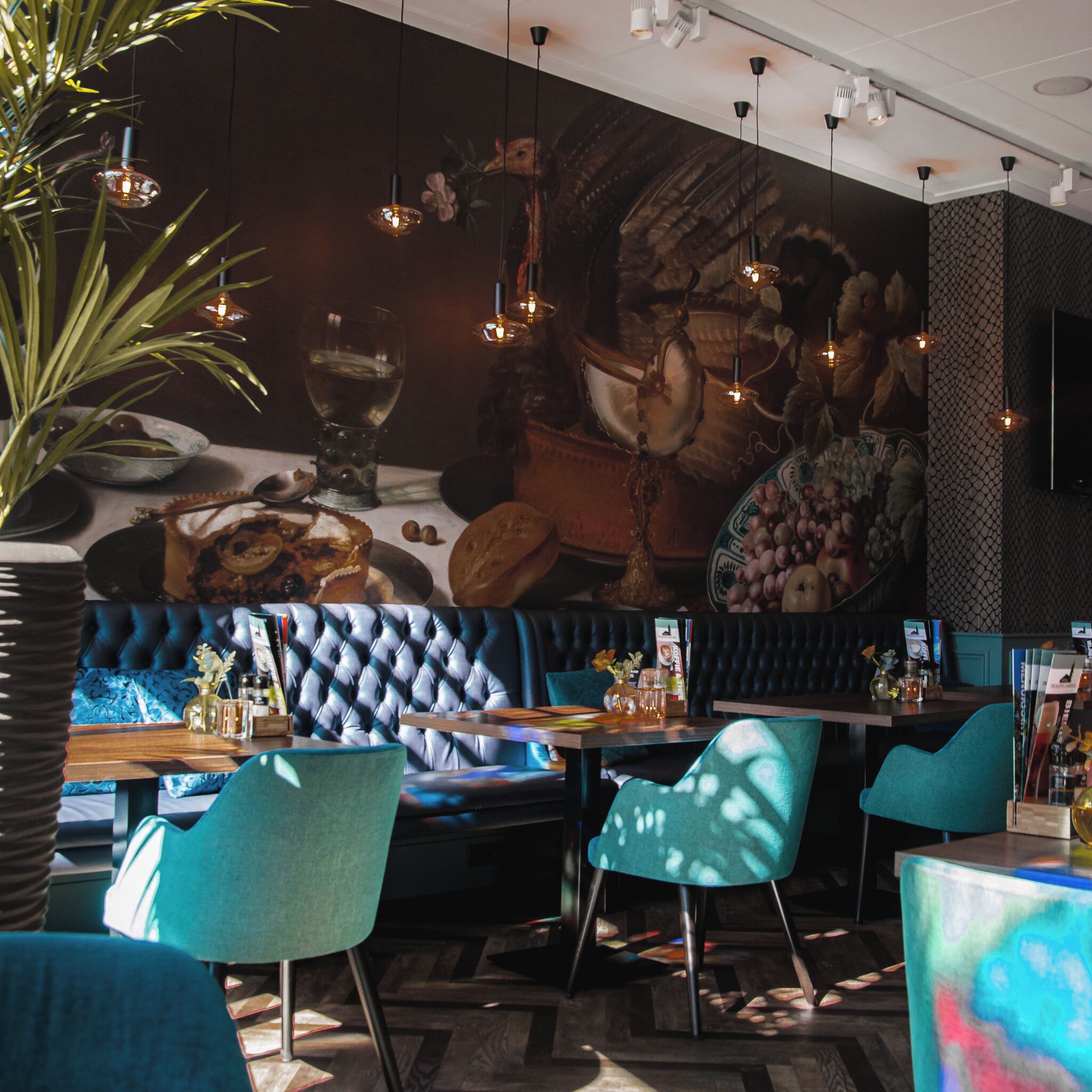 Un restaurant avec des banquettes bleues rembourrées, des chaises turquoise et une fresque représentant des fruits et des légumes sur le mur.