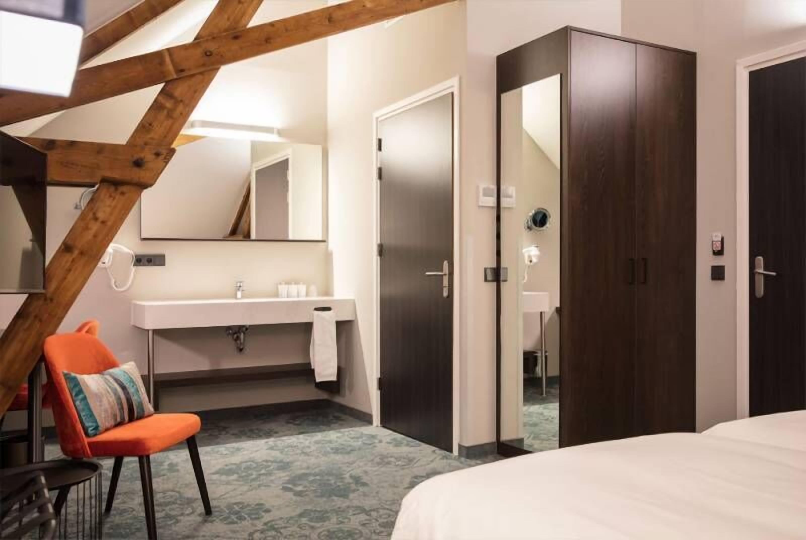 A hotel room with a wooden ceiling, a white bed, a bathroom with a sink and mirror, and a dark wardrobe.