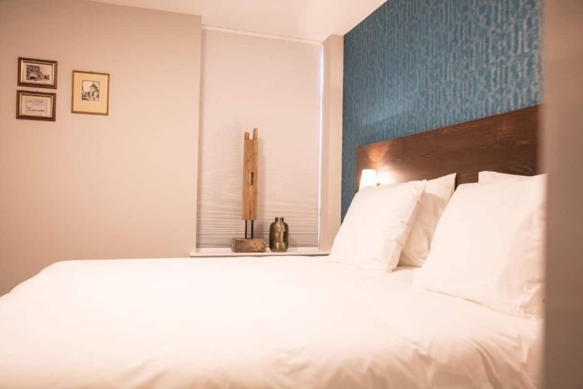 A hotel room with a large bed, a wooden headboard, a window with blinds, and two framed pictures on the wall.