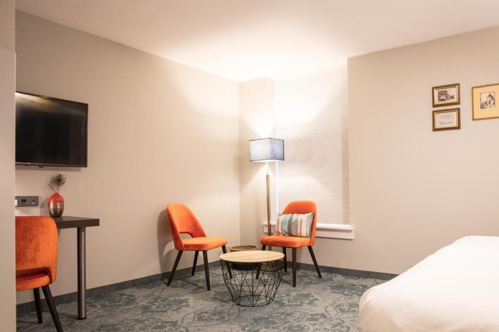 Ein Hotelzimmer mit einem Bett, zwei orangefarbenen Sesseln, einem kleinen runden Tisch, einem Fernseher, einer Lampe und gerahmten Bildern an der Wand.