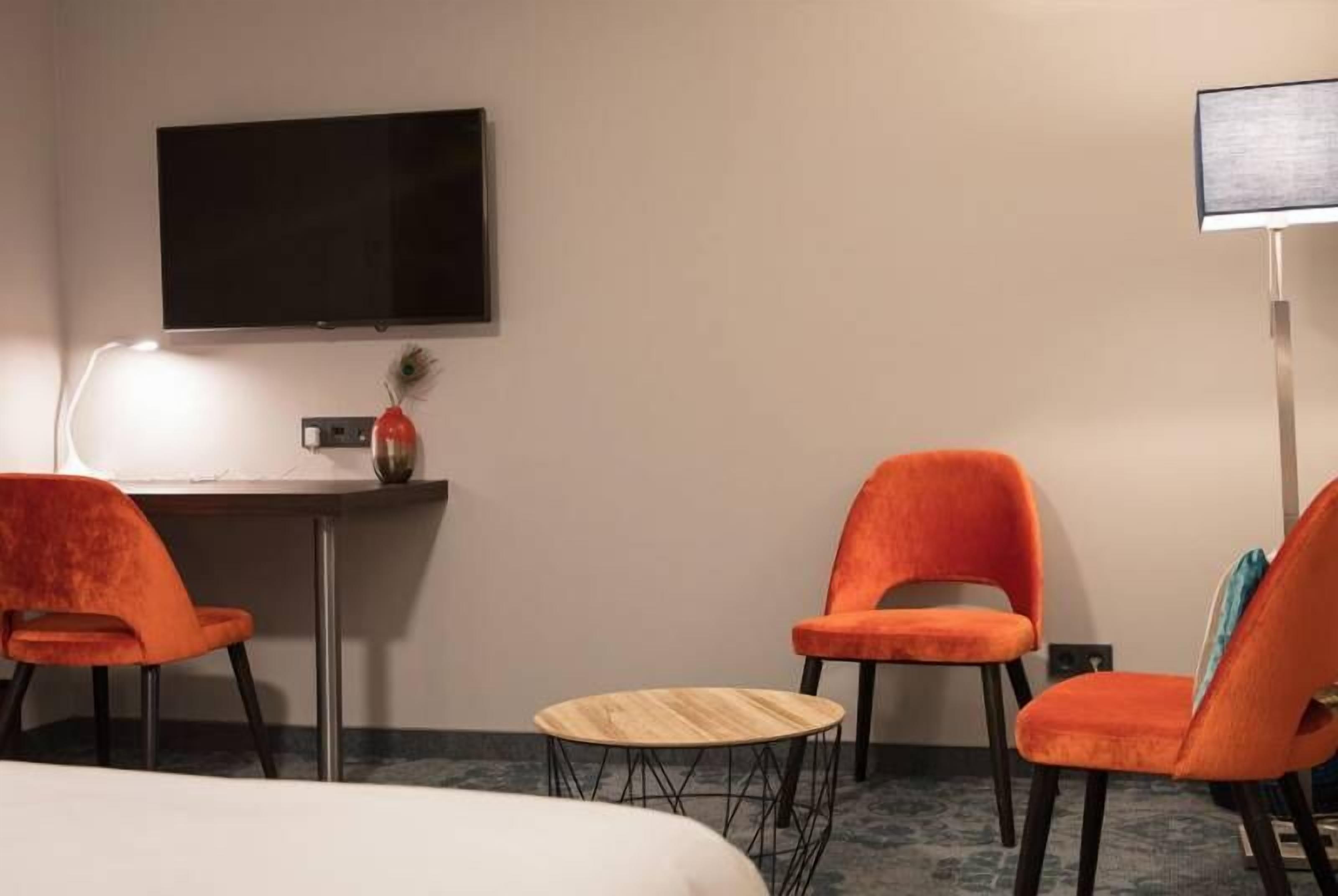 Habitació d'hotel amb un llit, dues cadires taronges, una taula de fusta petita, un televisor de pantalla plana fixat a la paret i un escriptori amb una làmpada i un objecte decoratiu.
