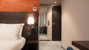 A hotel room with a wooden headboard, a bedside table with a lamp, a phone, and a small shelf. There is a door leading to a bathroom and a hallway. - Hotel de Koppelpaarden (Lichtenvoorde)
