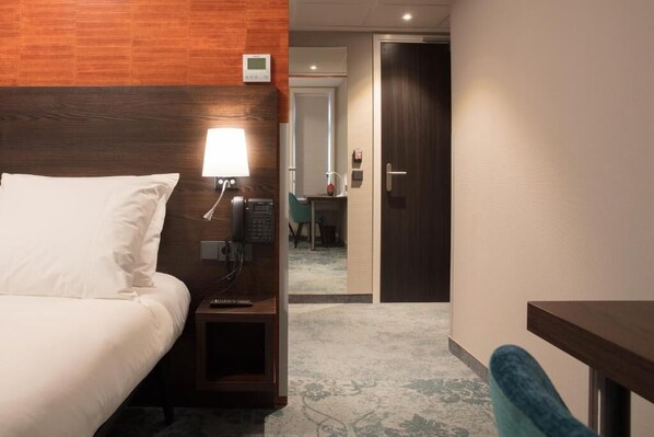 Habitación de hotel con cabecera de madera, mesita de noche con lámpara, teléfono y estante pequeño. Se ve una puerta que da al baño y a un pasillo.