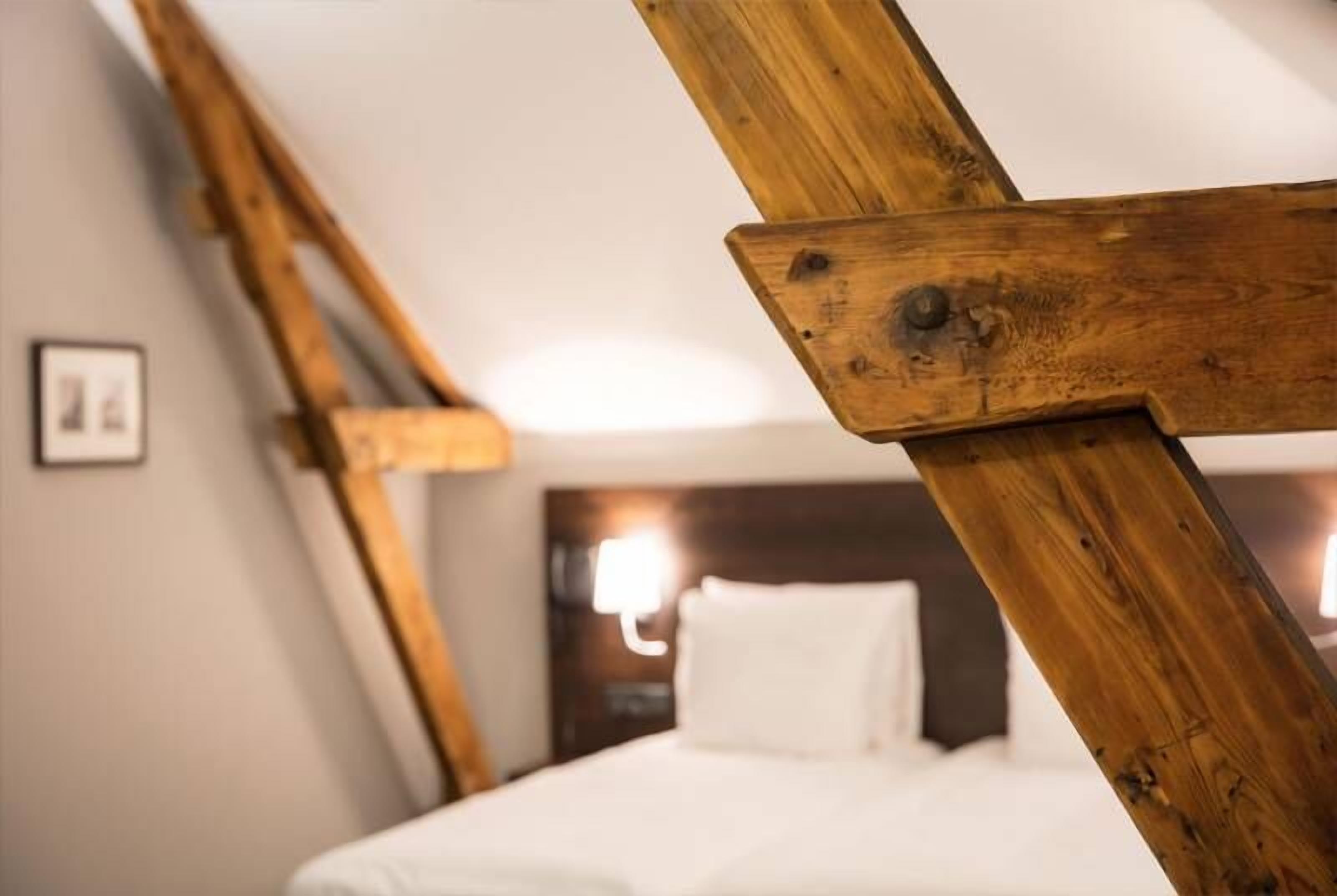 A room with a sloped ceiling, wooden beams, a bed with white bedding, and a wall-mounted light fixture.