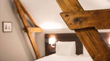 A room with a sloped ceiling, wooden beams, a bed with white bedding, and a wall-mounted light fixture.