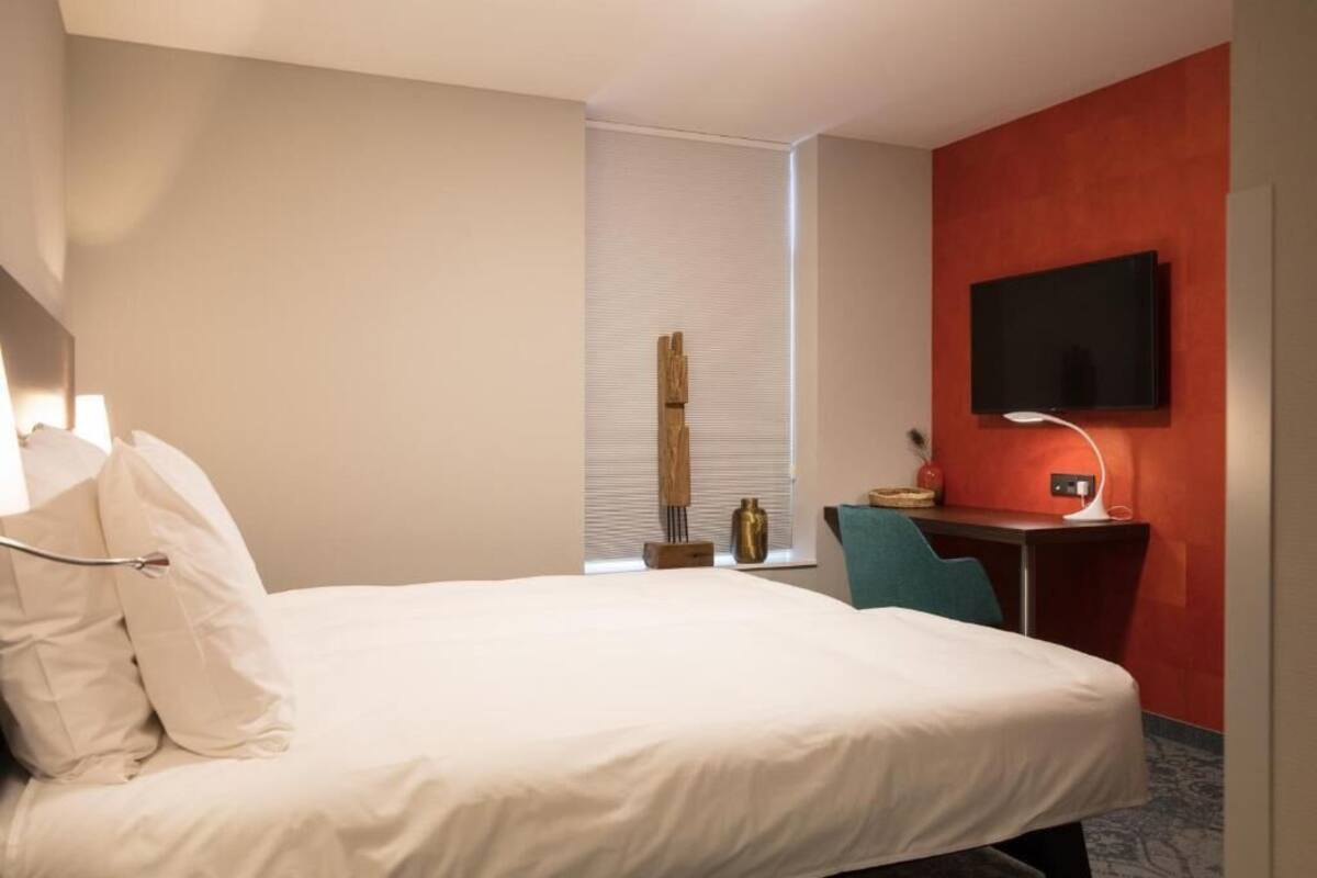 A hotel room with a bed, a desk with a computer, a television mounted on the wall, and a window with blinds.