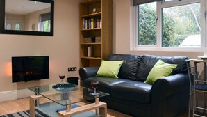 Uma sala de estar com sofá de couro preto, uma mesa de centro de vidro, estante de livros, TV de tela plana e janela com vista para árvores.